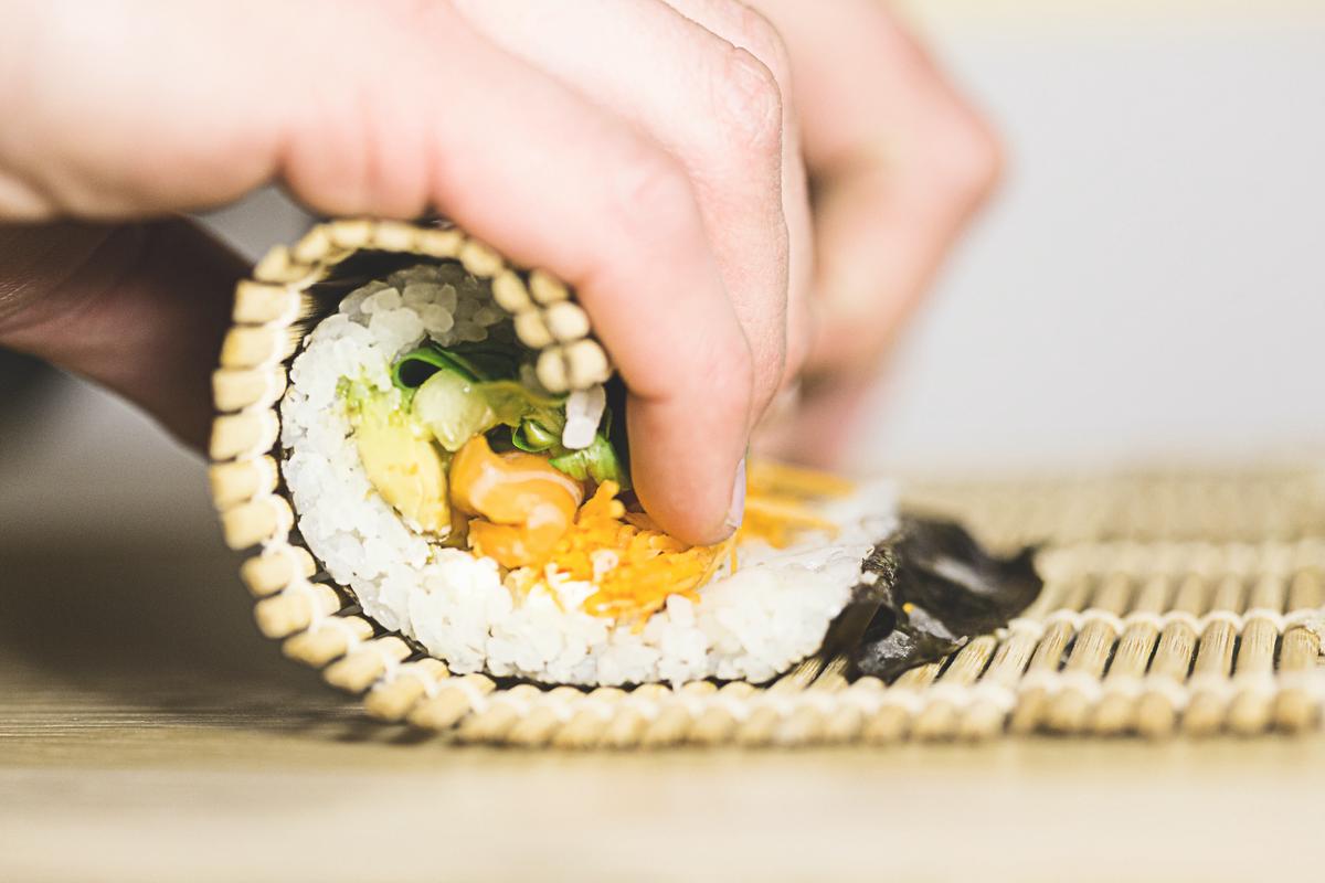 Bild mit einer visuellen Beschreibung: Eine Person rollt Sushi mit einer Bambusmatte und füllt es mit verschiedenen Zutaten. Es ist eine kunstvolle und präzise Methode, Sushi zuzubereiten.
